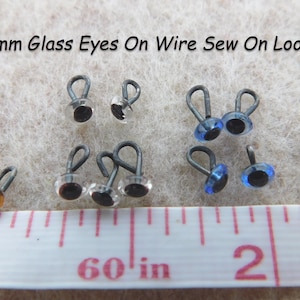 May include: Assortment of 3mm glass eyes with wire sew-on loops in brown, clear, and blue colors. The eyes are displayed on a white surface with a ruler measuring 1 to 2 inches.