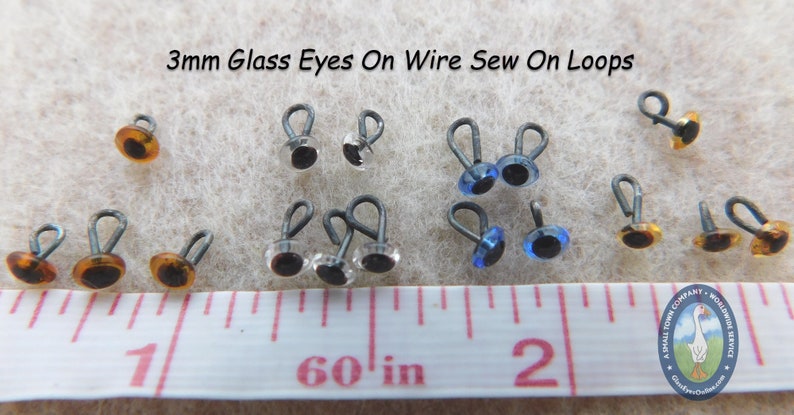 May include: Assortment of 3mm glass eyes with wire sew-on loops in brown, clear, and blue colors. The eyes are displayed on a white surface with a ruler measuring 1 to 2 inches.