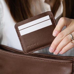 May include: A brown leather cardholder with two white card slots is held near a brown purse. The cardholder has a textured surface and a small logo in the corner. The purse is also brown.