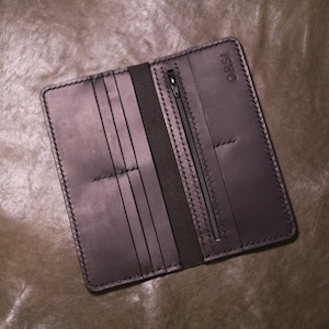 May include: Open, dark brown leather wallet with multiple card slots and a zippered pocket. The wallet is stitched with a contrasting thread and features the embossed word "ORSI" on the side. The wallet is displayed on a textured, brown surface.