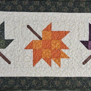 May include: A rectangular quilted table runner featuring three pieced fabric maple leaves in dark green, orange, and purple, set against a white background with stippled quilting. The runner has a dark green border with a floral pattern.