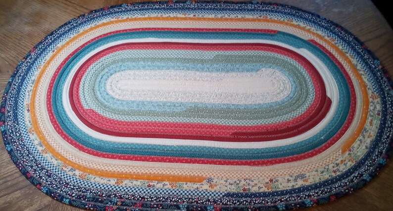 May include: A colorful oval braided rug with a white center and multiple bands of red, blue, yellow, and green fabric.