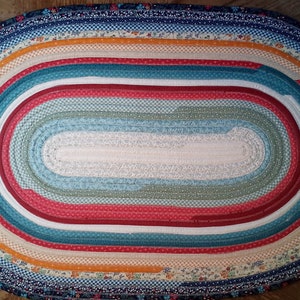 May include: Oval braided rug with a multi-colored design. The rug is made of fabric strips in shades of blue, green, red, white, and orange. The center of the rug is a solid white color.