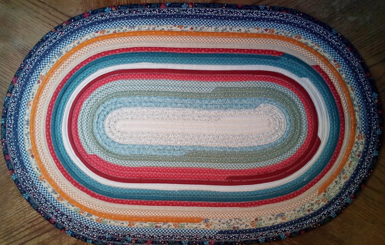 May include: Oval braided rug with a multi-colored design. The rug is made of fabric strips in shades of blue, green, red, white, and orange. The center of the rug is a solid white color.