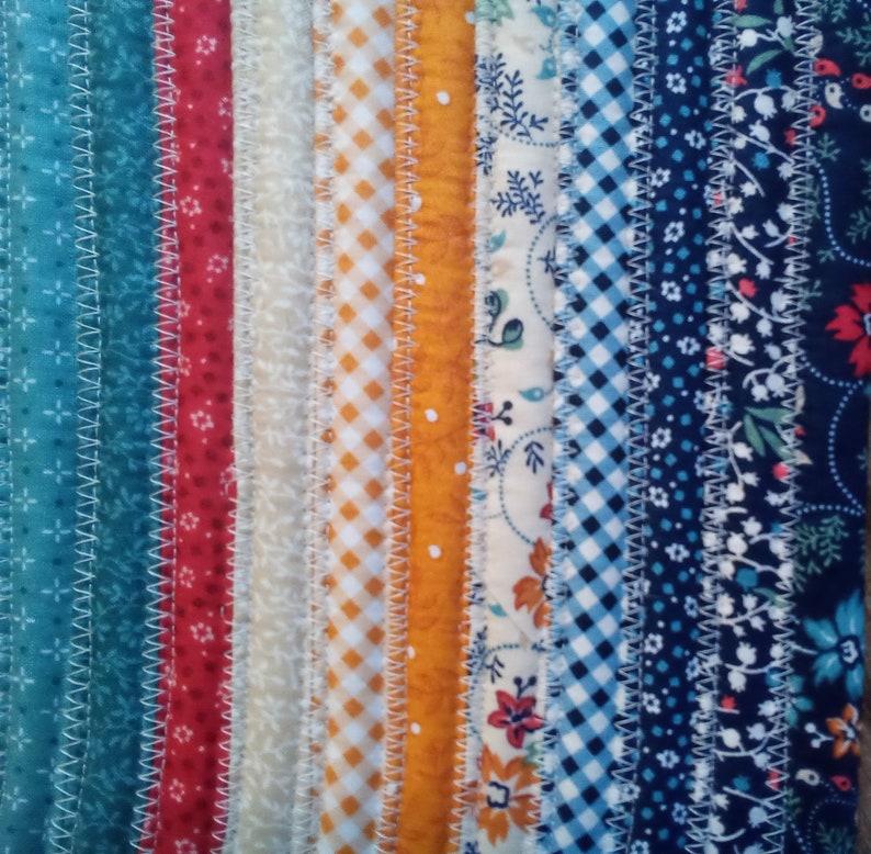 May include: A close-up of a colorful fabric strip quilt with various patterns, including floral, gingham, and polka dots. The fabric strips are sewn together with a white zig-zag stitch.