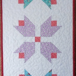 May include: A white quilted table runner with three rows of red, white, and blue fabric squares. Each row features a different pattern of fabric. The fabric squares are arranged in a starburst pattern.