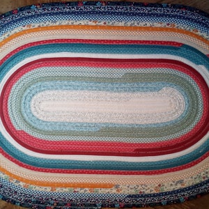 May include: Oval braided rug with multiple layers of fabric in shades of blue, red, white, green, and orange. The rug is made of strips of fabric sewn together to create a rope-like texture.