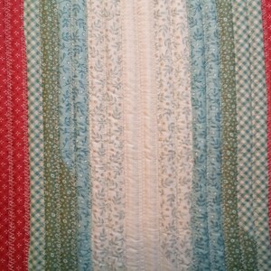 May include: A close-up of a quilt with a variety of fabric strips in red, green, blue, and white. The strips are sewn together in a vertical pattern, creating a striped effect. The quilt is made with a variety of fabrics, including floral prints, gingham, and solid colors.