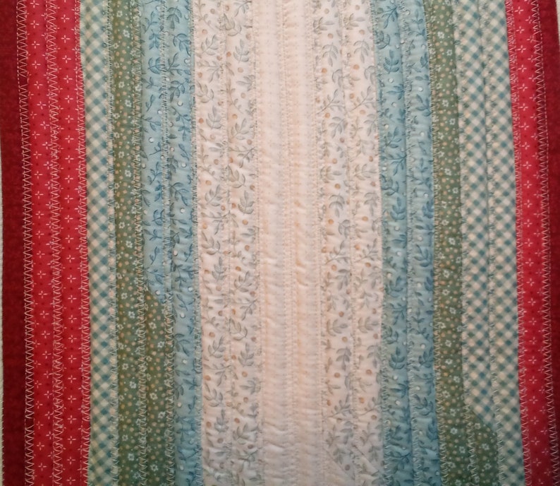 May include: A close-up of a quilt with a variety of fabric strips in red, green, blue, and white. The strips are sewn together in a vertical pattern, creating a striped effect. The quilt is made with a variety of fabrics, including floral prints, gingham, and solid colors.