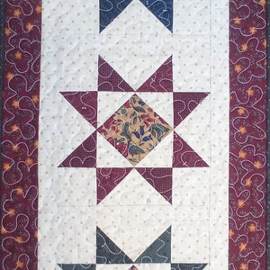 May include: A white quilted table runner with three star-shaped blocks. Each block features a different patterned fabric in shades of brown, beige, green, and red. The runner has a burgundy border with a floral pattern.