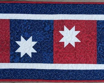 May include: A rectangular quilted table runner with a patriotic theme. The runner features alternating red and blue squares, each with a white star. A white border surrounds the central design, and a blue border with a red edge completes the piece.