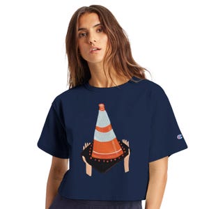 Pu&ograve; includere: Top corto Champion blu navy con una grafica di un cono stradale tenuto da mani. Il cono &egrave; arancione, bianco e grigio, con una base nera. Il logo Champion &egrave; sulla manica sinistra.