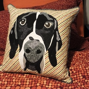 Op de afbeelding: Een decoratief kussen met een zwart-wit hondenhoofd op een gestreepte achtergrond. Het kussen is gemaakt van stof en heeft een gestreept patroon in bruin, oranje en geel.
