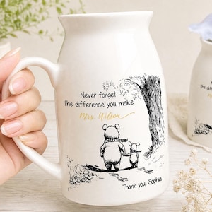 May include: A white ceramic mug featuring a black ink illustration of two characters walking near a tree. The text reads, "Never forget the difference you make," followed by "Mrs. Wilson" in gold script and "Thank you, Sophia" in black text.