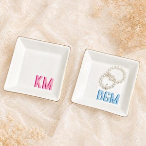 May include: Two square white ceramic trinket dishes with gold-toned rims. One dish features the pink letters KM, and the other dish features the blue letters BGM with a small white pearl-like beaded bracelet resting inside the tray.