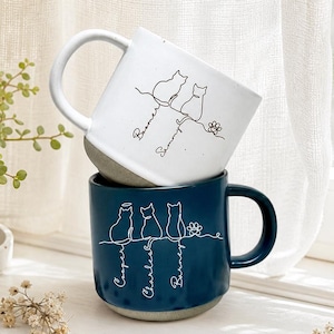 Peut inclure: Un mug en céramique blanc avec un dessin au trait noir de deux chats et les noms Boomer et Sammy, empilé sur un mug en céramique bleu foncé avec un dessin au trait blanc de trois chats et les noms Cooper, Charles et Barney, avec une patte.