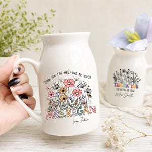 May include: Two white ceramic mugs with a flared rim and handle. The front features a colorful floral illustration with the text "THANK YOU FOR HELPING ME GROW" and the name "MRS. REGAN" in a retro font, followed by "Love Julia".