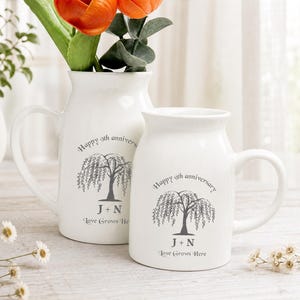 May include: Two white ceramic pitchers with handles, each featuring a gray printed design of a willow tree, the text "Happy 9th anniversary", the initials "J + N", and the phrase "Love Grows Here" printed on the front surface of each vessel.
