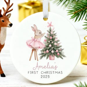 Puede incluir: Adorno navideño redondo con una ilustración en acuarela de un conejito con tutú rosa decorando un árbol de Navidad. El adorno tiene el texto "Amelia's First Christmas 2025". Se adjunta una cinta blanca para colgarlo.
