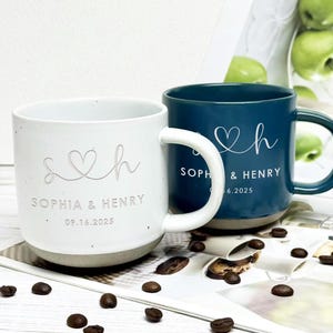 Könnte beinhalten: Zwei Keramikbecher, einer weiß und einer blaugrün, mit den Namen "Sophia & Henry" und einem Datum. Der weiße Becher hat eine gesprenkelte Oberfläche und einen grauen Boden. Der blaugrüne Becher hat einen passenden Henkel. Kaffeebohnen sind verstreut.