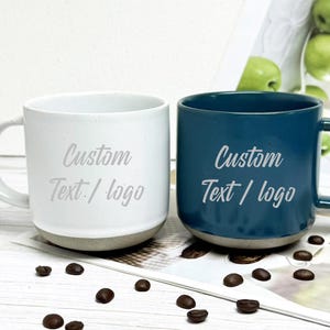 May include: Two ceramic mugs, one white and one teal, with a gray base and handle. The text "Custom Text / logo" is printed on the front of each mug in a light gray color. Coffee beans are scattered around the mugs.