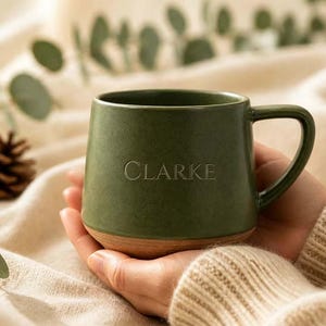 May include: A matte olive green ceramic mug with a rounded shape and a single handle. The name CLARKE is engraved in a simple serif font on the front of the mug. The base of the mug features an exposed, unglazed natural brown clay texture.