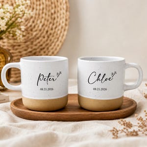 Puede incluir: Dos tazas de cerámica blanca con textura moteada y base de color canela. Cada taza presenta texto en cursiva negra con un pequeño icono de corazón. La taza izquierda muestra el nombre Peter y la fecha 08.21.2026, la derecha el nombre Chloe y la fecha 08.21.2026.