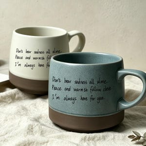 Può includere: Due tazze in ceramica con manico, una color crema e una blu-grigio. Entrambe presentano una base marrone e il testo stampato in nero: Don't bear sadness all alone. Peace and warmth follow close. I'm always here for you. Finitura liscia.