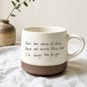 May include: A two-toned ceramic mug with a cream-colored upper section and a dark brown base. The cream section features black printed text that reads: Don't bear sadness all alone. Peace and warmth follow close. I'm always here for you.