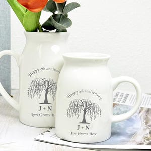 May include: Two white ceramic mugs with handles, one holding flowers. Each mug features a weeping willow tree design with the text "Happy 9th anniversary" and "Love Grows Here" along with initials. The mugs are on a light background.