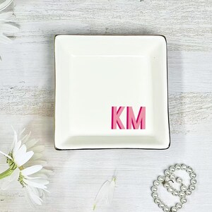 以下が含まれることがあります： 黒い縁取りと中央にピンク色の文字「KM」が入った正方形の白いセラミック製の小物入れ。白い木製の表面に置かれ、白い花とパールのブレスレットが背景にあります。