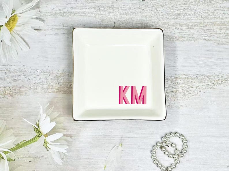 以下が含まれることがあります： 黒い縁取りと中央にピンク色の文字「KM」が入った正方形の白いセラミック製の小物入れ。白い木製の表面に置かれ、白い花とパールのブレスレットが背景にあります。
