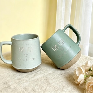 May include: Two ceramic mugs with speckled finishes in light green and sage green. Each mug features an engraved tally mark design, a small heart symbol, and the text "Jack & Lisa". The mugs have a smooth glazed upper section and a textured base.