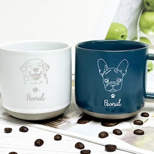 May include: Two ceramic mugs, one white and one teal, each with a dog illustration and the name "Peanut." The white mug has a golden retriever design, while the teal mug showcases a French bulldog. Both mugs have a light grey base.