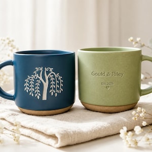 Puede incluir: Dos tazas de cerámica con acabado mate. La taza izquierda es azul oscuro y presenta un sauce llorón grabado en blanco. La taza derecha es verde salvia y tiene el texto grabado Gould & Riley, Est.2026 y un pequeño corazón. Ambas tienen una base color canela.