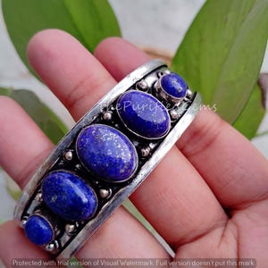 May include: A silver cuff bracelet featuring five oval and round, deep blue lapis lazuli stones. The stones are set in a detailed, ornate silver band. The Purified Gems is printed on the bracelet.