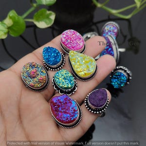 以下が含まれることがあります： ピンク、青、黄色、紫など、さまざまな色の宝石リングのコレクション。各リングは、銀色の金属バンドにセットされたユニークな質感の石が特徴です。リングは手の上に表示されています。