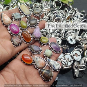 Puede incluir: Una colección de anillos de piedras preciosas con varios colores y formas, incluyendo piedras ovaladas, en forma de lágrima y redondas. Los anillos tienen un engaste de metal plateado con bordes decorativos. El texto "ThePurifiedGems" es visible.