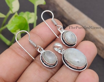 Puede incluir: Un conjunto de joyas de plata con pendientes y un colgante. Cada pieza tiene una piedra ovalada de color claro engastada en un marco plateado con un borde negro. Los pendientes tienen un diseño en espiral.