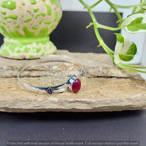 May include: A silver bangle bracelet featuring a large, oval red gemstone. The bracelet has a delicate design with small heart-shaped details. The gemstone is set in a silver bezel. The bracelet is displayed on a textured, light brown surface.