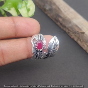 Op de afbeelding: Zilverkleurige ring met een veermotief en een ronde, rode edelsteen. De ring wikkelt zich om de vinger met twee veervormige details aan weerszijden van de edelsteen. The Purified Gems.