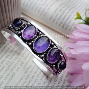Può includere: Un bracciale a polsino in argento con pietre preziose ovali e rotonde di colore viola. Il bracciale ha un design ornato con pietre incastonate. Le pietre preziose sono di un colore viola intenso e il metallo ha una finitura antica.
