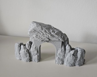 Może przedstawiać: Szary miniaturowy łuk skalny o teksturowanej powierzchni. Łuk posiada kamienny łuk i jest otoczony formacjami skalnymi. Model został zaprojektowany tak, aby przypominał naturalną formację skalną.