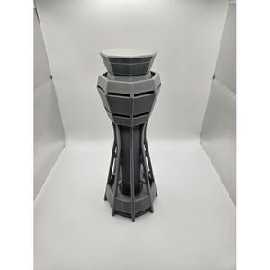 May include: A 3D-printed model of an airport control tower. The tower is primarily gray with a lighter gray top. The design features a detailed structure with multiple levels and a cylindrical base. The model is a miniature representation of an airport control tower.