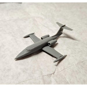 May include: A gray model airplane with two engines, wings, and a tail fin. The miniature jet is a detailed replica, likely 3D-printed, and sits on a textured, off-white surface. The plane's design suggests a business jet or private aircraft.