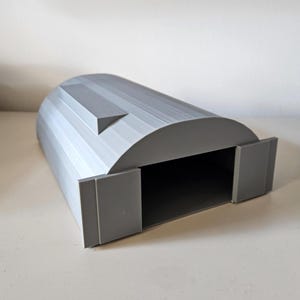 May include: A gray, 3D-printed model of a Quonset hut. The structure has a curved roof with vertical ridges and a rectangular opening with two small doors. The model is made of a solid, light gray material.