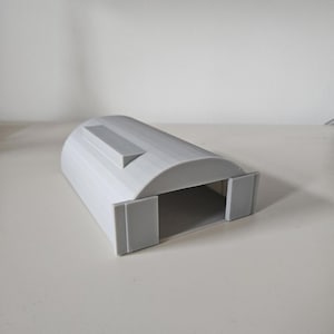 May include: A light gray, 3D-printed model of a small arched building or hangar. The structure has a rectangular base with a curved roof and a small rectangular protrusion on top. Two rectangular panels form the entrance.