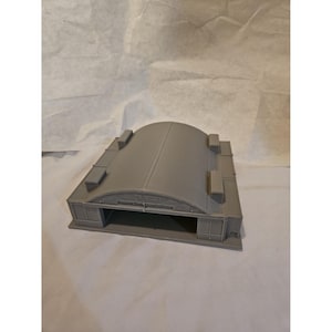 May include: A gray model of an aircraft hangar. The structure has a curved roof, rectangular side sections, and a large opening at the front. Four rectangular protrusions are on the roof.