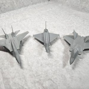 May include: Three gray model fighter jets are arranged on a white surface. The jets are different designs, with wings and tails. The models are detailed, showing the shape of the aircraft.