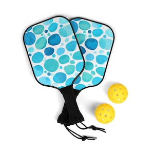 Puede incluir: Dos palas de pickleball con caras azules y blancas con patrones y mangos negros, y dos pelotas de pickleball amarillas.